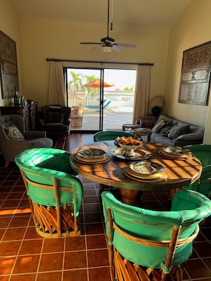 Un coin repas avec une table ronde, des chaises recouvertes d’un tissu vert et une vue sur l’extérieur grâce à une porte vitrée.