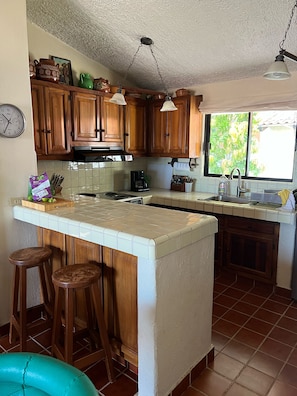 A kitchen with wooden cabinets, a central island with stools, a clock, and a window with a view of greenery. - Wonderful 2-bedroom house with AC in San José del Cabo (San José del Cabo)
