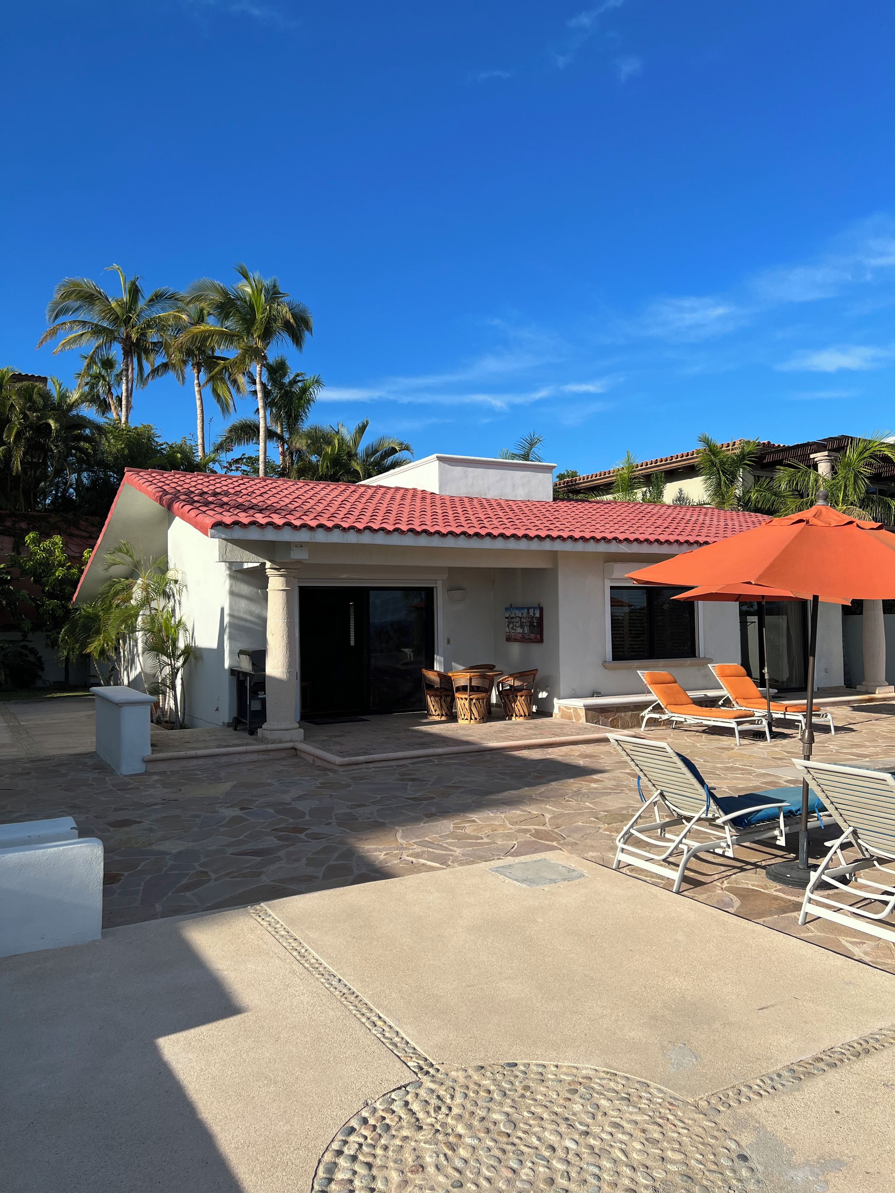 Un bâtiment avec un toit recouvert de tuiles rouges, un espace extérieur aménagé avec des chaises et des tables, et des palmiers en arrière-plan.