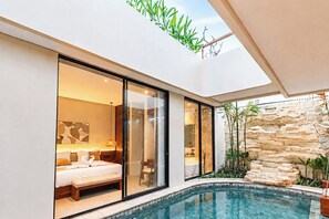 Una habitación de hotel moderna con pared de vidrio, cama, mesita de noche y baño visible a través del vidrio.