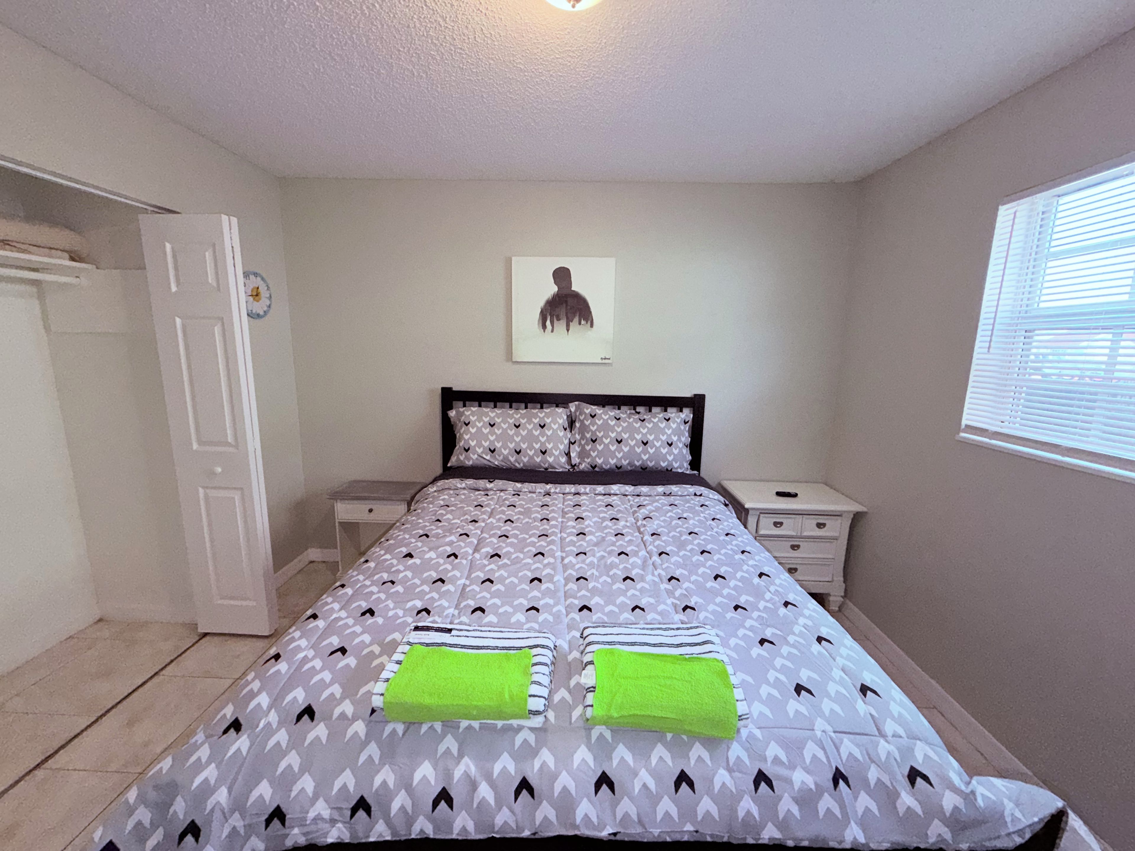 A bedroom with a bed, two bedside tables, a door, a window with blinds, and a wall-mounted picture.
