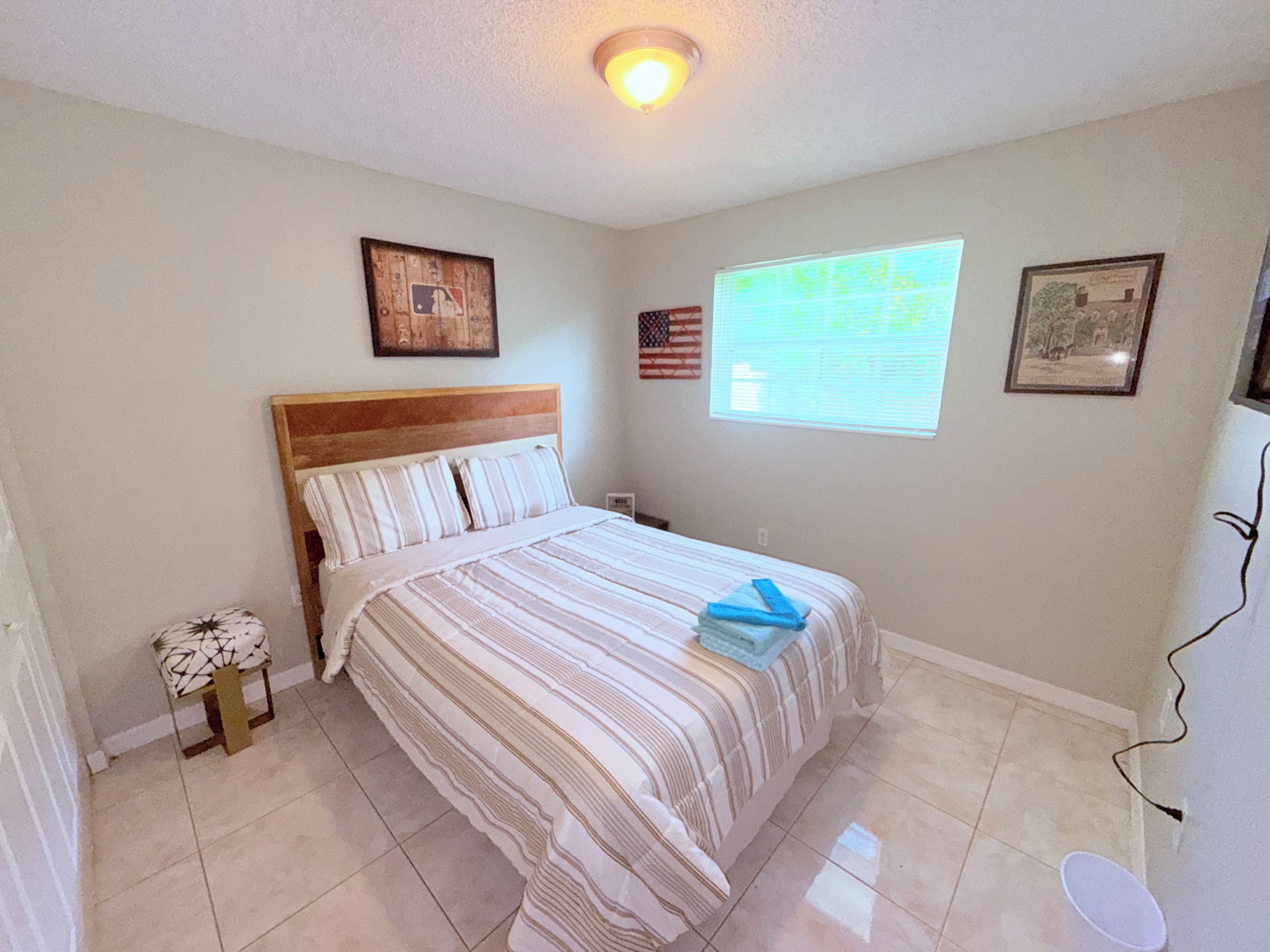A bedroom with a bed, a small table, a window with blinds, and a ceiling light.