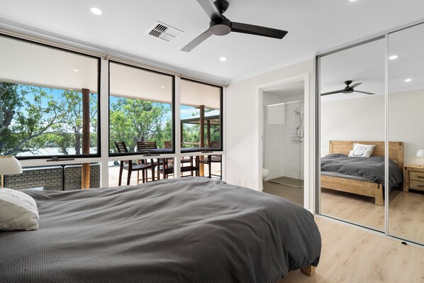 A modern bedroom with a large bed, a ceiling fan, and a view of the outdoors through large windows.
