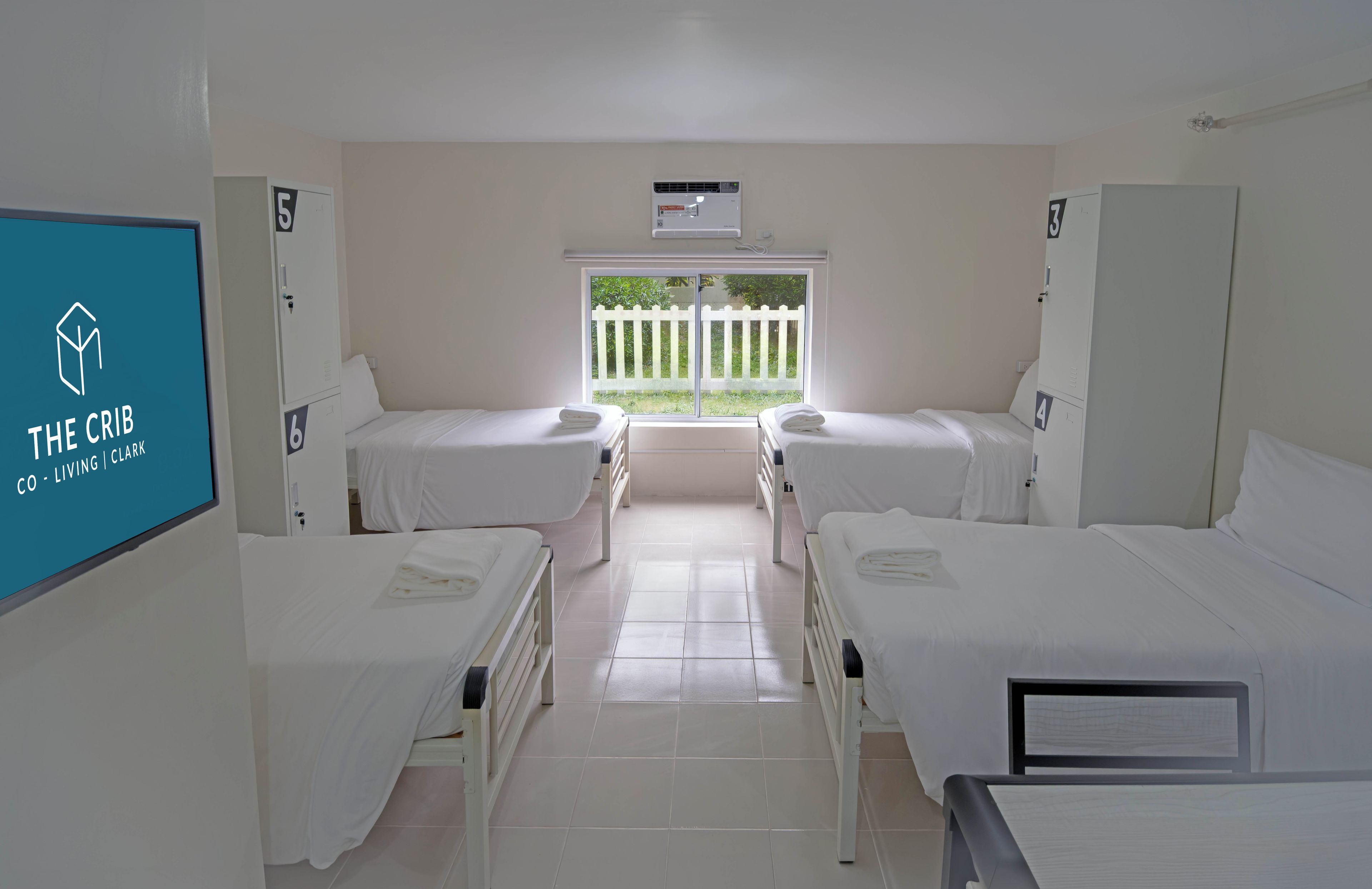 Habitación de hotel con dos camas individuales, una ventana con vista a una valla blanca y un aire acondicionado instalado.