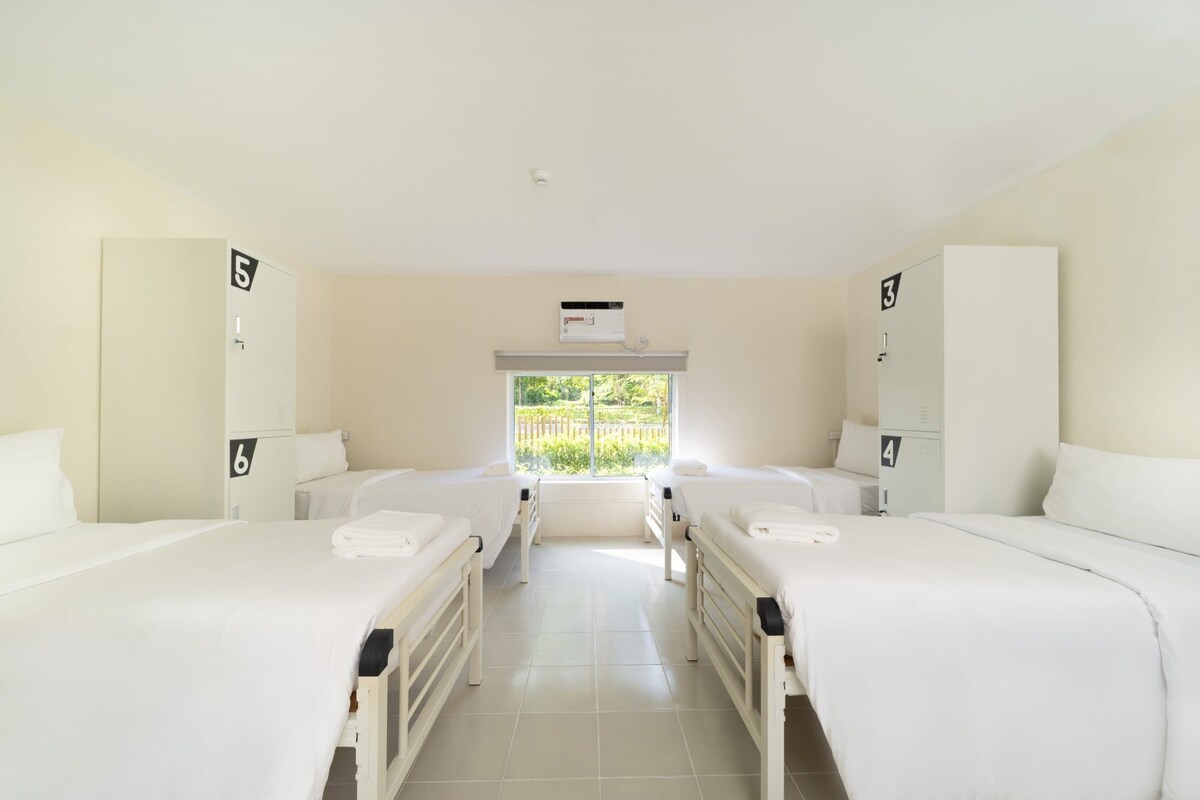A hotel room with two beds, a window with a view of greenery, and a wall-mounted air conditioning unit.