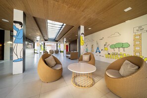 A spacious lobby with high ceilings, a skylight, and a mural depicting people in various activities. There are wicker chairs and a round table in the center. - The Crib Clark (Mabalacat City)