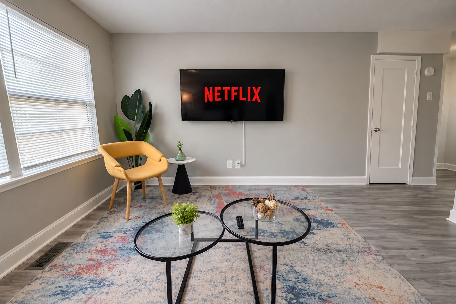 A living room with a flat-screen TV mounted on the wall, a yellow chair, a small table with a plant, and a coffee table with decorative items.