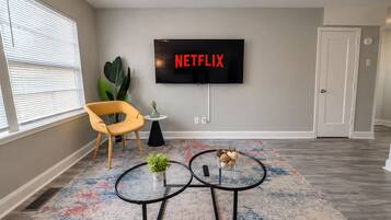 A living room with a flat-screen TV mounted on the wall, a yellow chair, a small table with a plant, and a coffee table with decorative items.