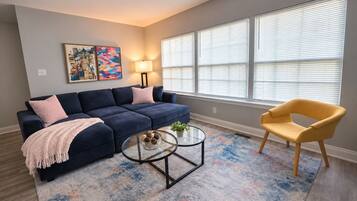Un salon avec un canapé bleu foncé, un fauteuil jaune, une table basse en verre et un tapis à motifs.