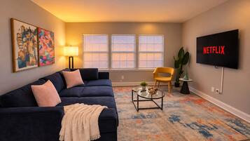 A living room with a dark blue sofa, a yellow armchair, a glass coffee table, and a flat-screen TV mounted on the wall.