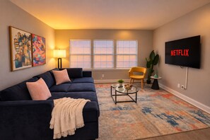 Una sala de estar con un sofá azul oscuro, un sillón amarillo, una mesa de centro de vidrio y un televisor de pantalla plana montado en la pared.