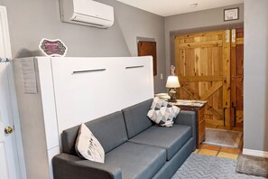 Ein Zimmer mit einer grauen Couch, einem weißen Kühlschrank, einer Holztür und einem kleinen Tisch mit einer Lampe.