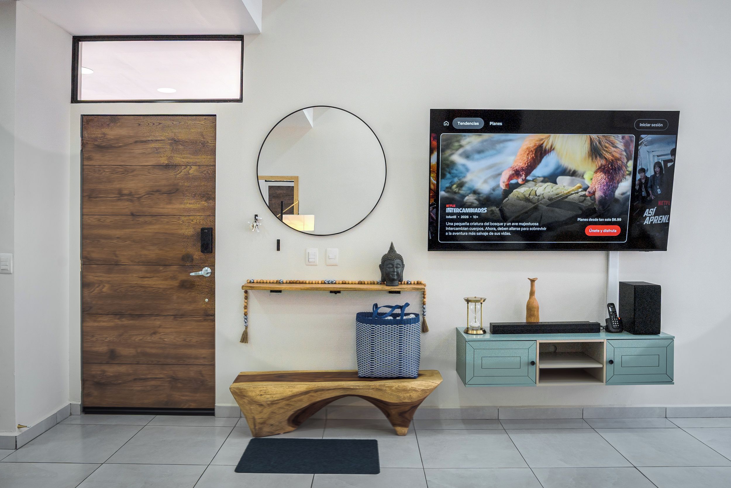 A modern living room with a wooden door, a round mirror, a wooden bench, a television mounted on the wall, and a small shelf with decorative items.