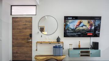 A modern living room with a wooden door, a round mirror, a wooden bench, a television mounted on the wall, and a small shelf with decorative items.