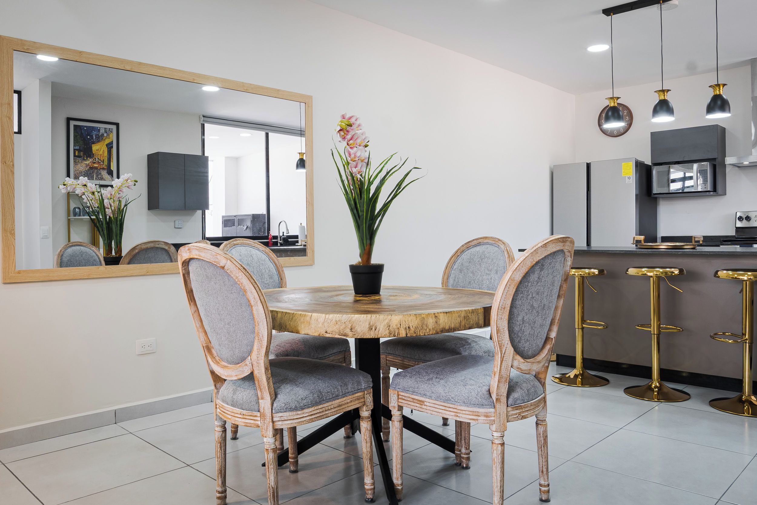 Een eetruimte met een ronde houten tafel, vier gestoffeerde stoelen, een potplant en een grote spiegel aan de muur.