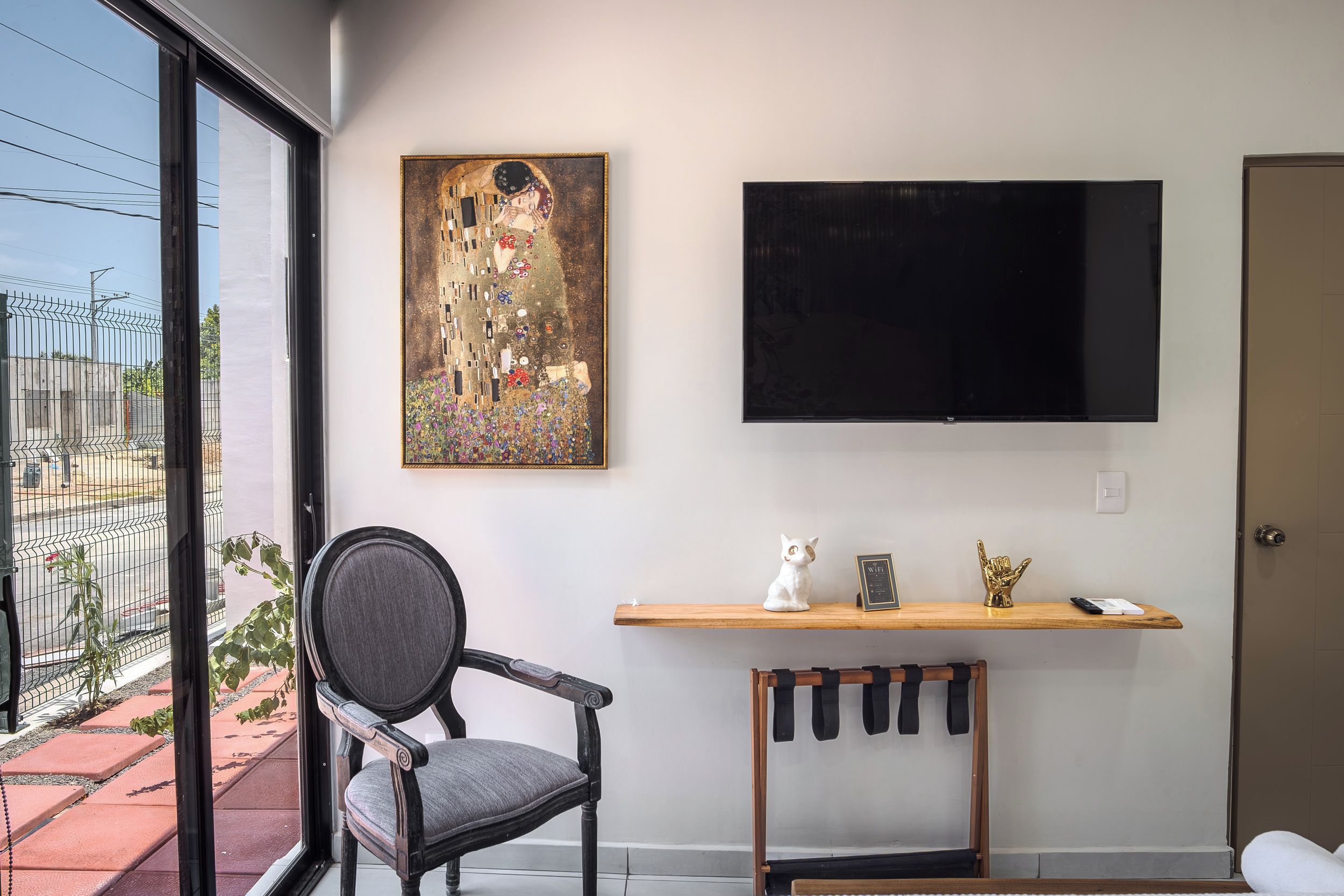 Een kamer met een groot raam, een schilderij aan de muur, een televisie bevestigd boven een houten plank en een stoel.