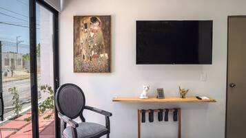 Een kamer met een groot raam, een schilderij aan de muur, een televisie bevestigd boven een houten plank en een stoel.