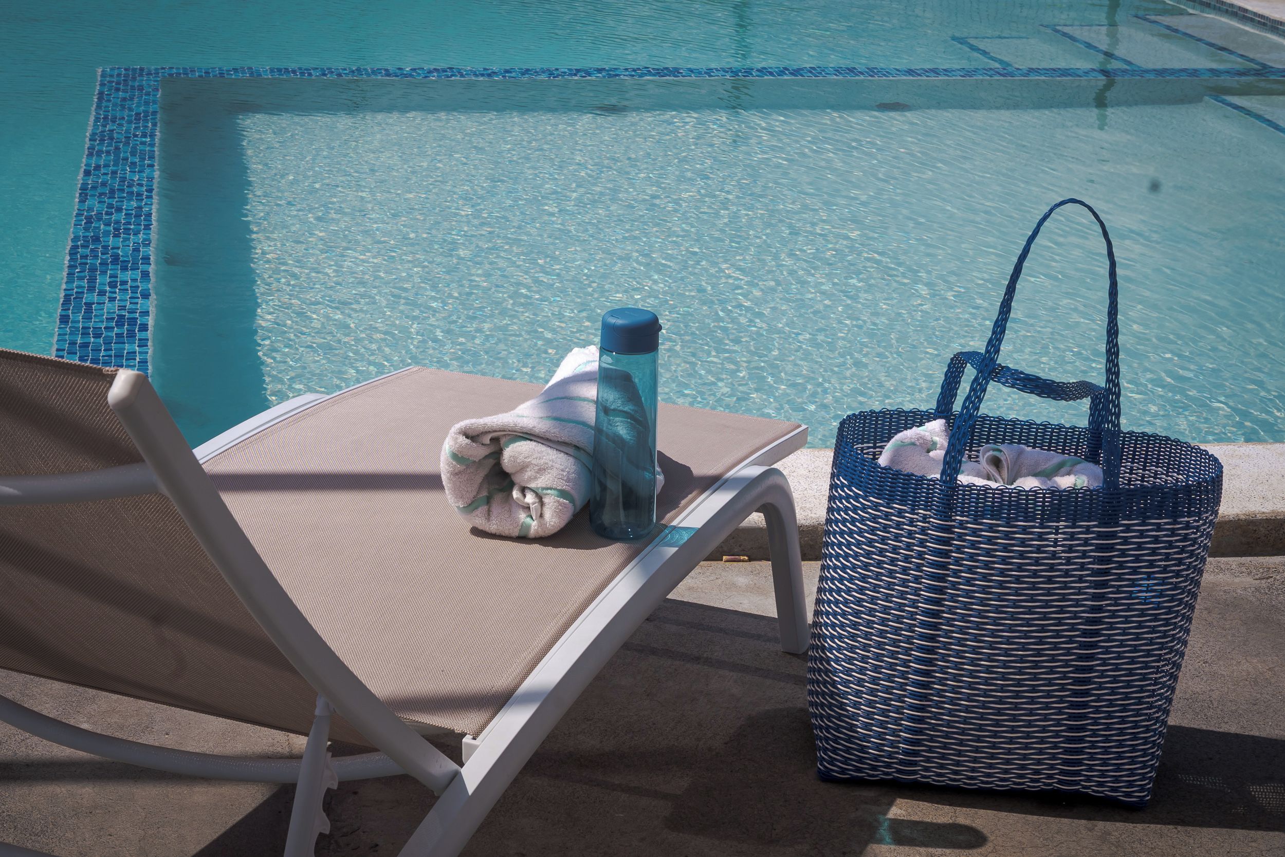 A poolside area with a lounge chair, a towel, a water bottle, and a woven bag.
