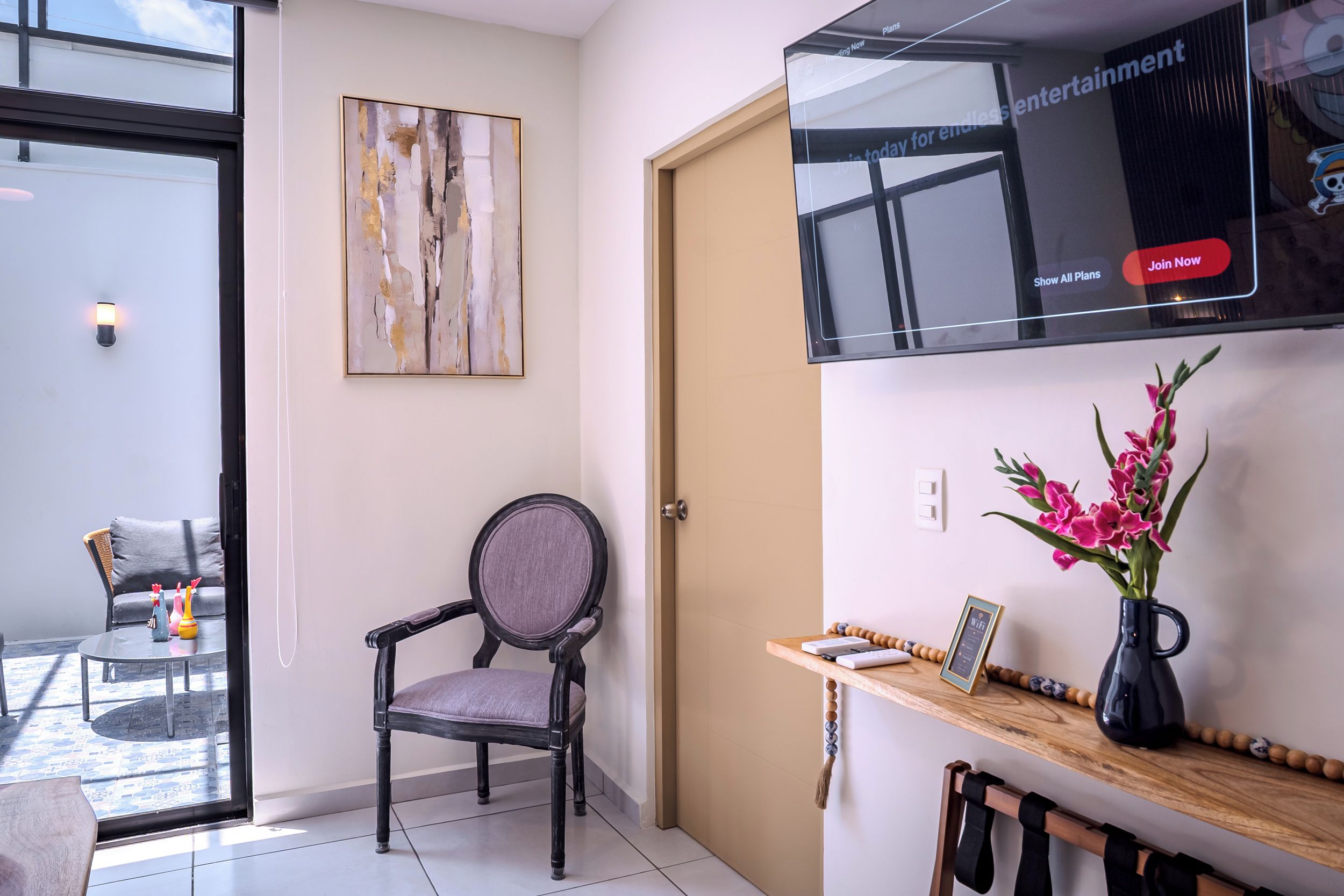 Ett modernt vardagsrum med en glasdörr som leder ut till en balkong, en mörk trästol, ett litet bord med en vas med blommor och en väggmonterad TV som visar en skylt.