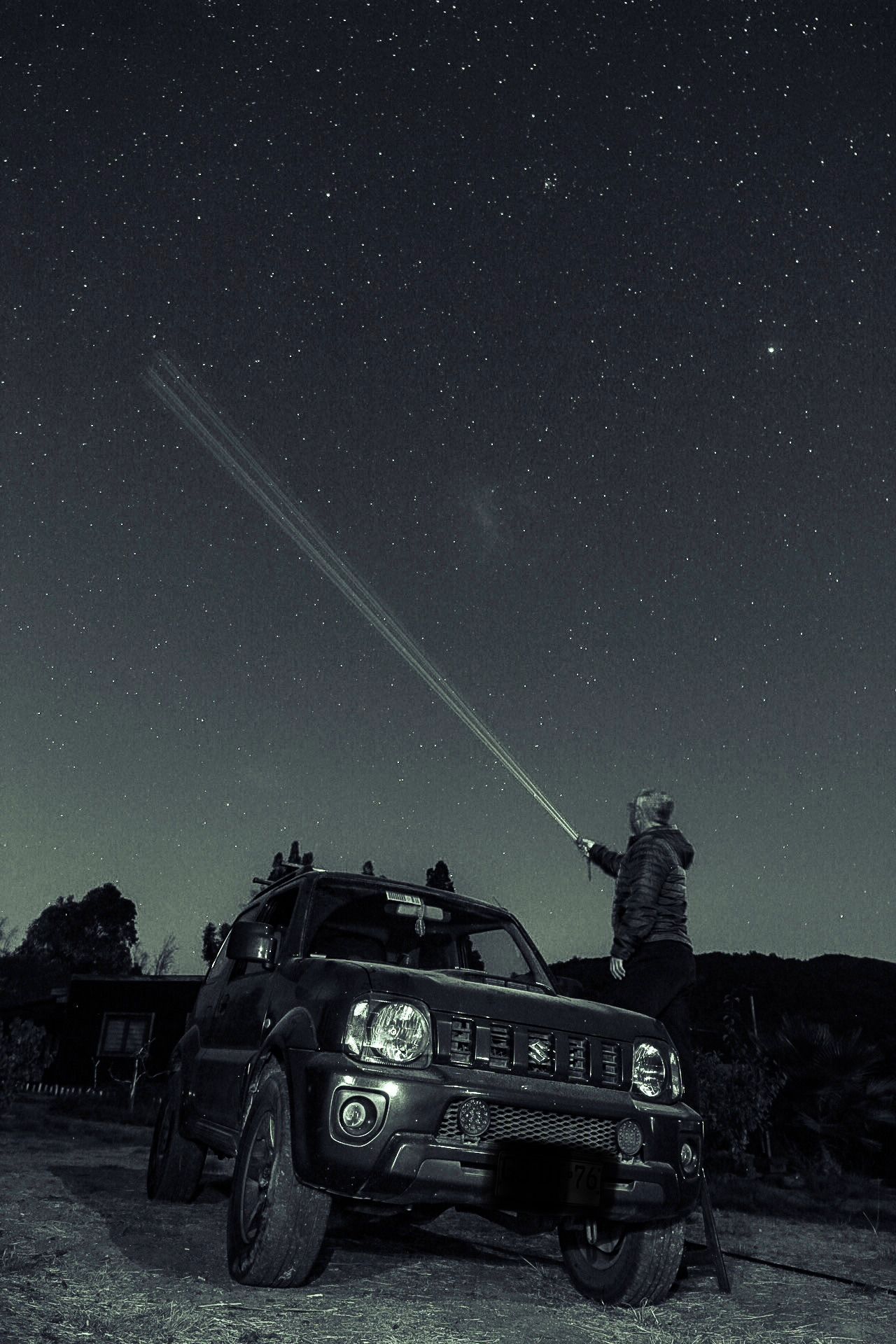 Un coche antiguo estacionado afuera de noche, con una persona sosteniendo un objeto largo, posiblemente una luz o un láser, apuntando hacia el cielo.