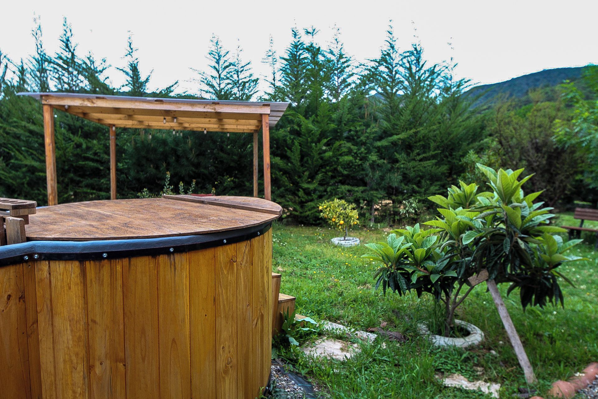 A wooden hot tub with a wooden deck and a pergola structure, surrounded by a lush garden with various plants and a bench in the background.
