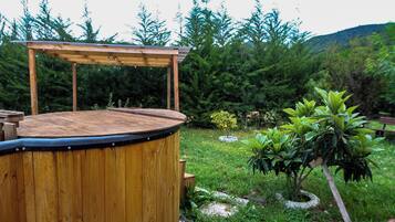 A wooden hot tub with a wooden deck and a pergola structure, surrounded by a lush garden with various plants and a bench in the background.