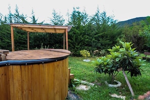 Un bain à remous en bois, doté d’une terrasse en bois et d’une structure en forme de pergola, entouré d’un jardin luxuriant avec différentes plantes et d’un banc à l’arrière-plan.
