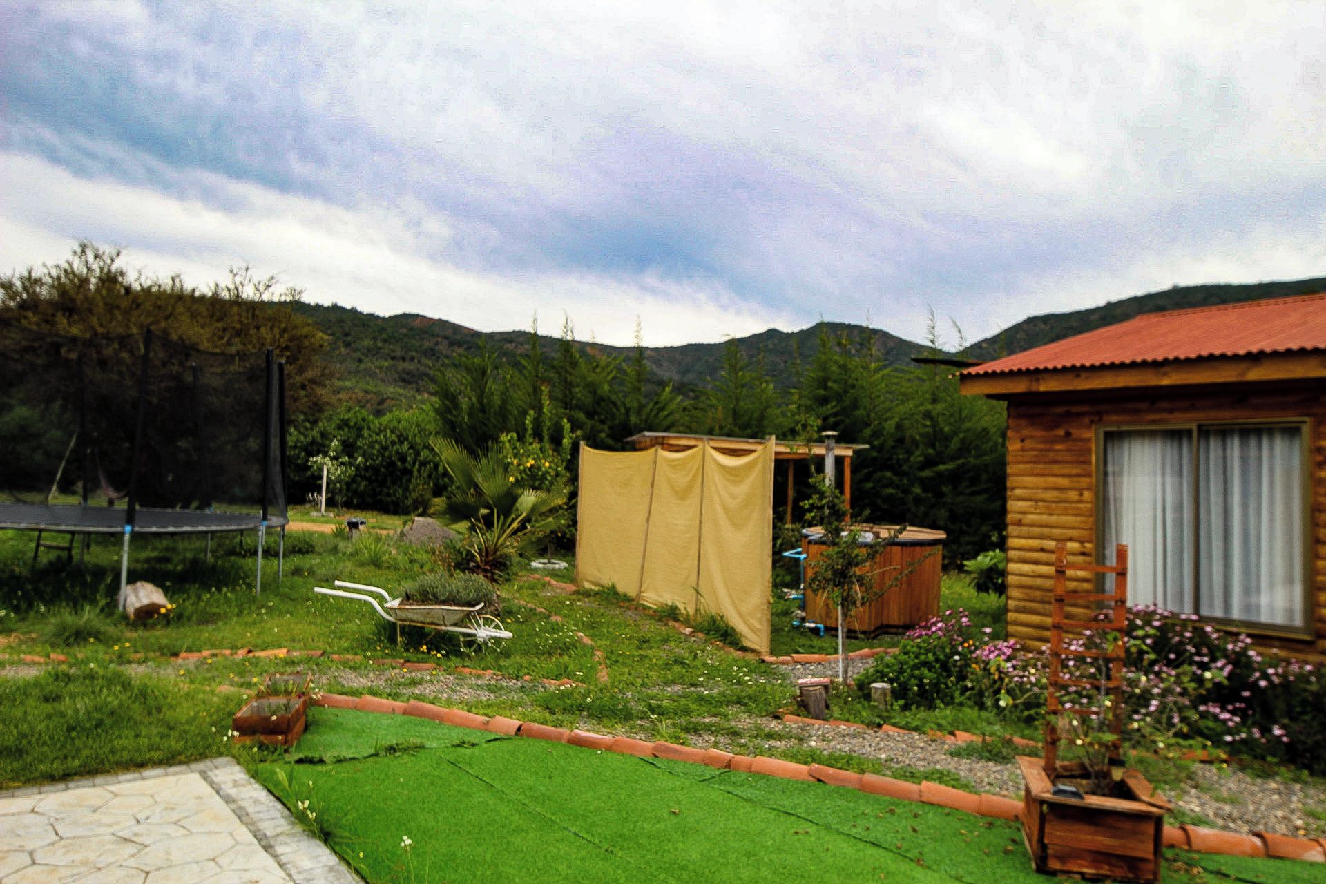 Una cabaña de madera con techo rojo, rodeada por un jardín con césped verde, un trampolín y una carretilla.