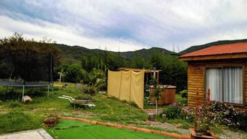 Una cabaña de madera con techo rojo, rodeada por un jardín con césped verde, un trampolín y una carretilla.