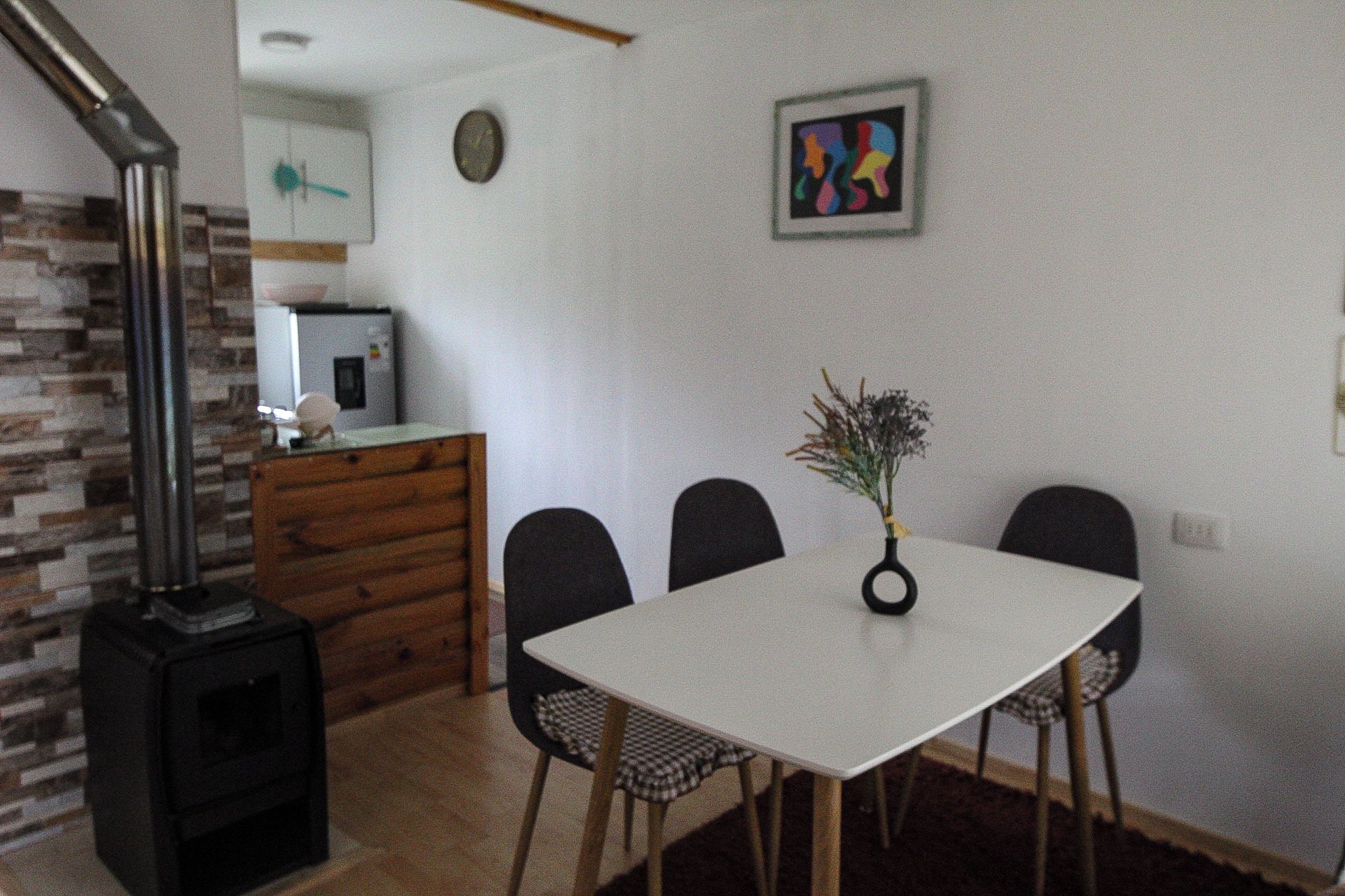 Uma sala de jantar com uma mesa branca, quatro cadeiras, um vaso com flores, um relógio e uma pintura abstrata colorida na parede.