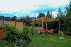 A covered outdoor seating area with two chairs and a table, surrounded by greenery and a tent in the background.