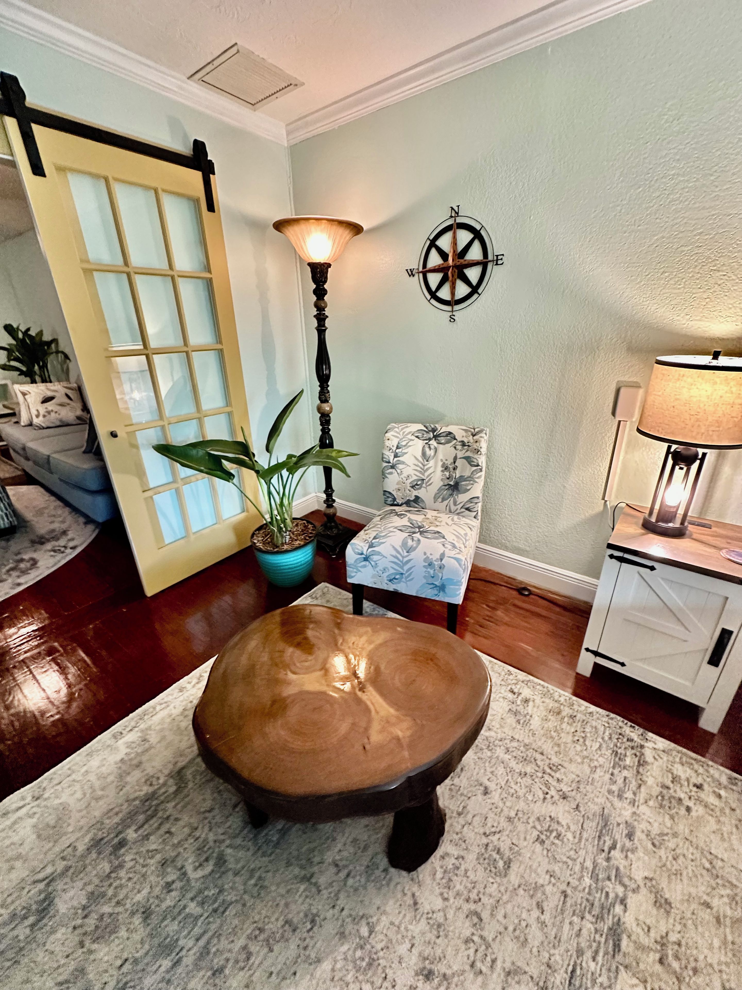 A room with a wooden bench, a potted plant, a chair with floral upholstery, a lamp, and a compass wall decoration.