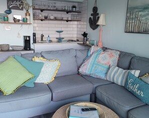 A living room with a grey sofa, decorative pillows, a coffee table with a book and remote, and a kitchen in the background. - END Condo directly on the Beach with unobstructed views of the ocean (Corpus Christi)