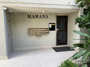 Ein Gebäudeeingang mit der Aufschrift 'MAHANA' über der Tür, einem Sicherheitstastaturfeld und einer Bank davor.