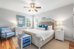 Une chambre à coucher comprenant un lit, une table de chevet, une chaise, une commode, un ventilateur de plafond et une fenêtre avec des stores.