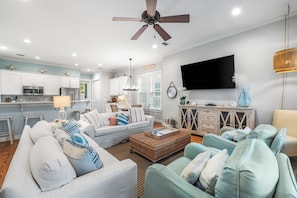 A modern living room with a large flat-screen TV, a ceiling fan, and a dining area with white cabinets and a kitchen in the background. - Salty Seagull - Private Beach Access, 4 bikes! (Seacrest)