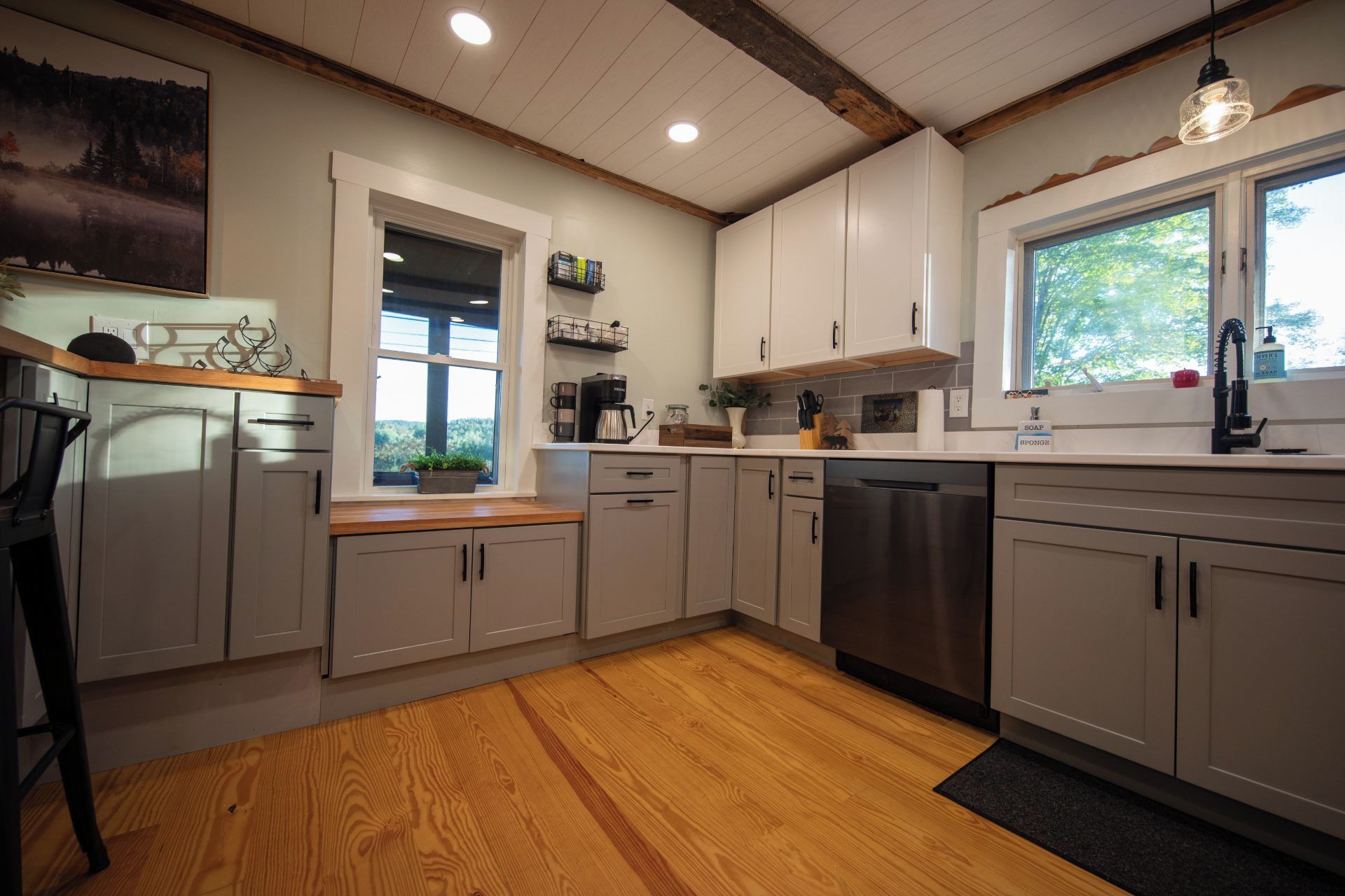 Una cocina moderna con suelo de madera, armarios blancos, lavavajillas integrado y ventana con vista a árboles.