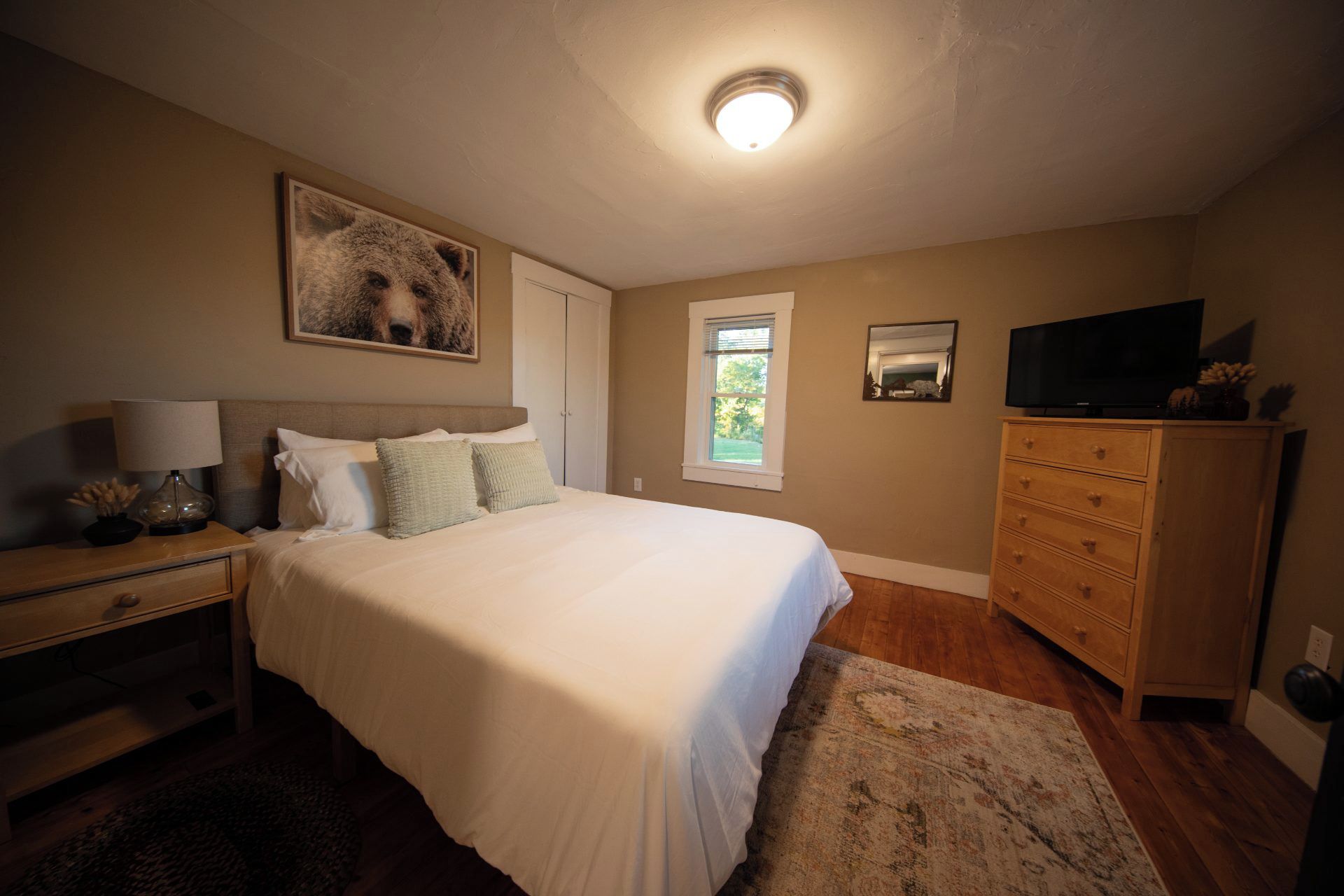 A bedroom with a large bed, a nightstand, a dresser, a television, and a window.