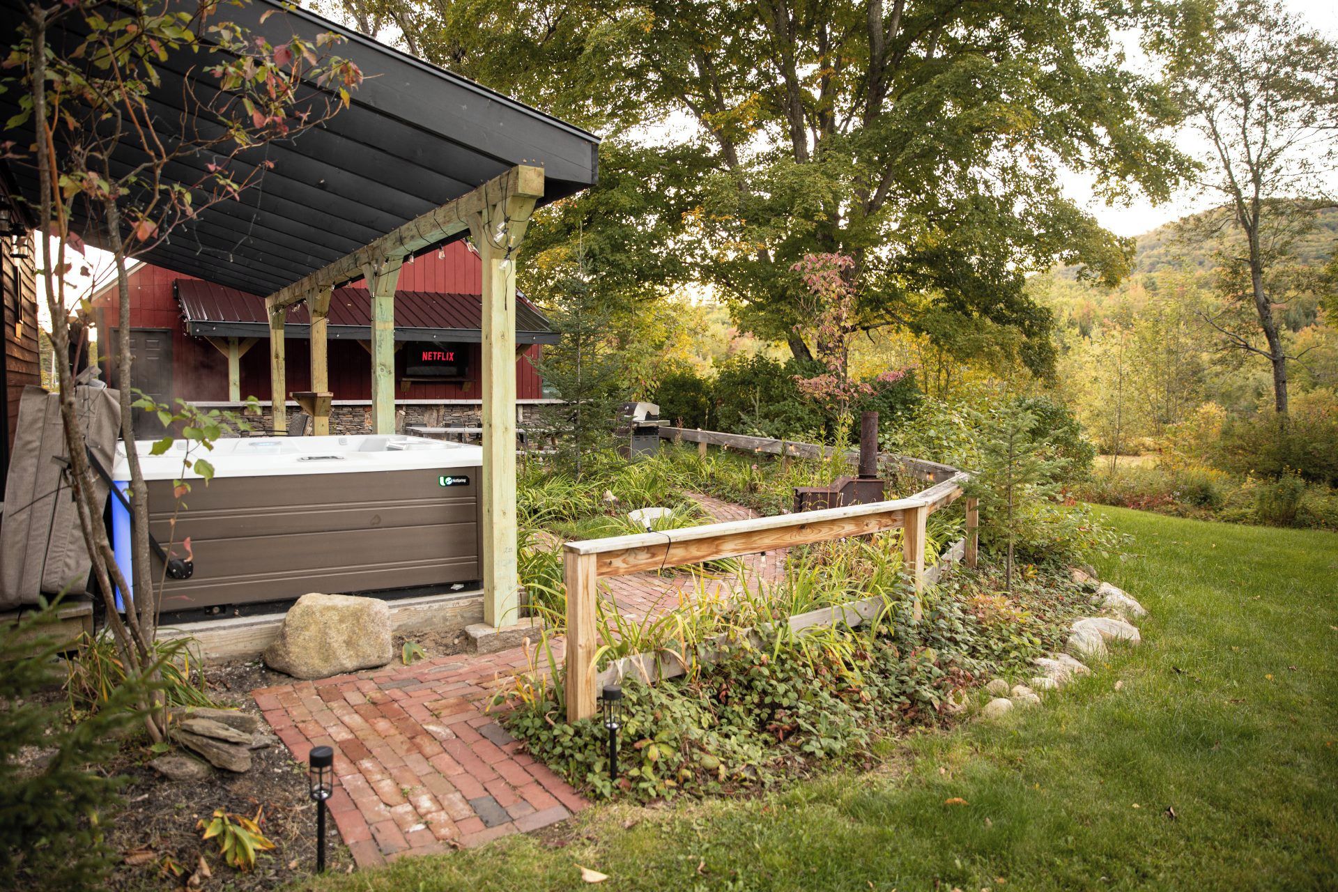Ein überdachter Außenbereich mit Whirlpool, umgeben von einem Garten mit Holzterrasse und Steinmauer.