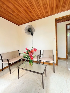 A room with a wooden ceiling, two sofas, a glass table with a vase of flowers, and a ceiling fan.