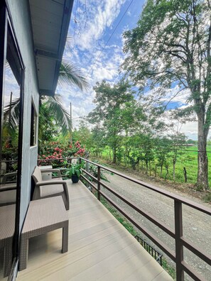 A balcony with wicker furniture, a view of a rural landscape with trees and a fence, and a clear sky.