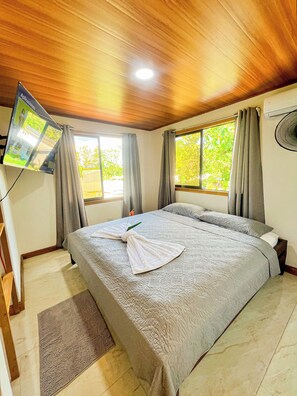 A hotel room with a bed, a flat-screen TV mounted on the wall, a window with curtains, and a ceiling fan.
