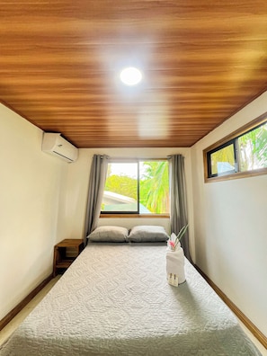 A bedroom with a wooden ceiling, a bed with a floral-patterned bedspread, a nightstand, an air conditioning unit, and a window with curtains.