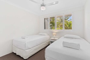 A room with two beds, a ceiling fan, a window with a view of greenery, and a nightstand with a lamp. - Miami Resort Living - Two Bedroom Resort Views (Gold Coast)