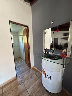 A bathroom with a glass-top sink, a mirror, and a white barrel with 'JM' on it. - Chácara JM: Your little corner to connect with nature and enjoy days of leisure. (Lindóia)