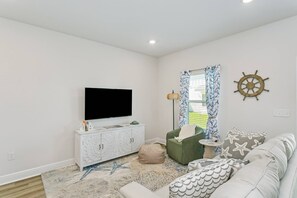 A living room with a flat-screen TV, a white cabinet, a green armchair, a patterned rug, and a wall-mounted ship's wheel. - Pool & Playground - Newly Built 3BR Gulf Breeze Townhome Near The Zoo, Navarre Beach, and Pensacola (Gulf Breeze)