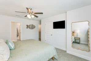 A bedroom with a bed, a ceiling fan, a flat-screen TV, a wall-mounted mirror, and a door leading to another room. - Pool & Playground - Newly Built 3BR Gulf Breeze Townhome Near The Zoo, Navarre Beach, and Pensacola (Gulf Breeze)