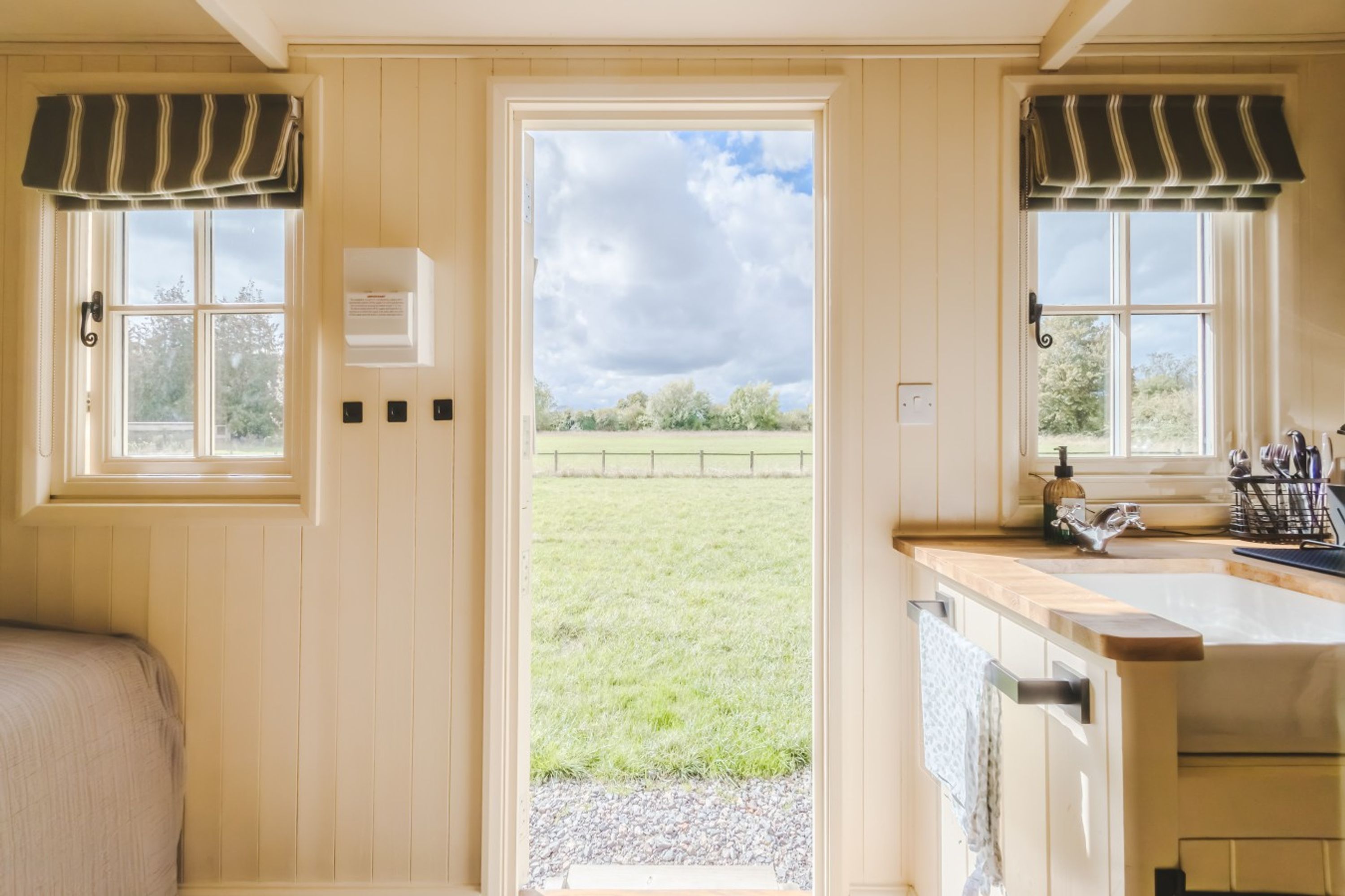 Una habitación con una puerta que da a un espacio exterior, un ventanal con vistas a un campo y una zona de cocina con pileta y utensilios.