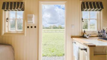 Una habitación con una puerta que da a un espacio exterior, un ventanal con vistas a un campo y una zona de cocina con pileta y utensilios.