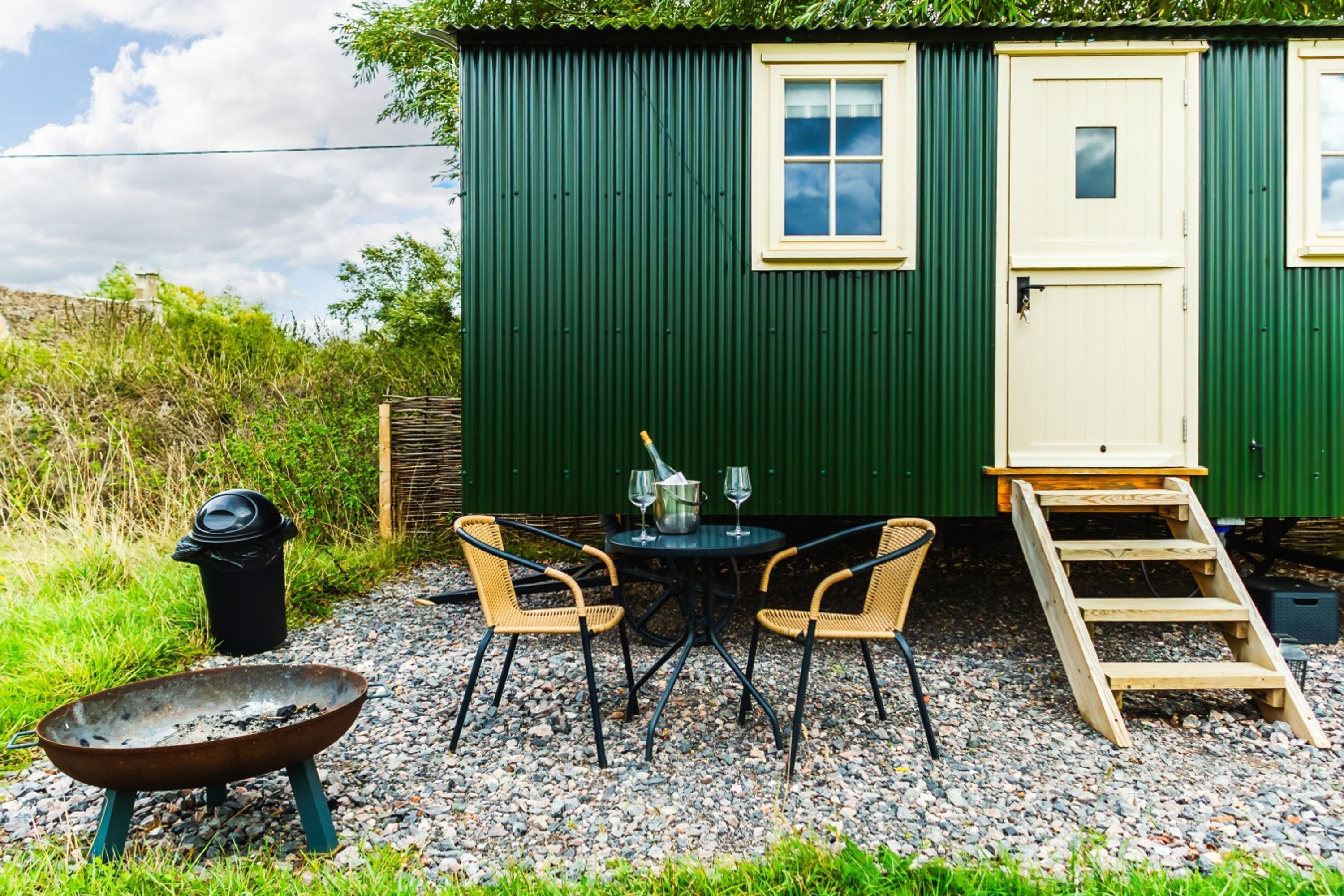 A small, green, single-room structure with a door, a window, and a table with chairs. There is a fire pit and a trash bin outside.